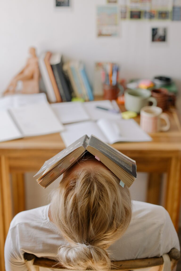 一個用書本蓋著頭很苦惱坐在書桌前的金髮女生